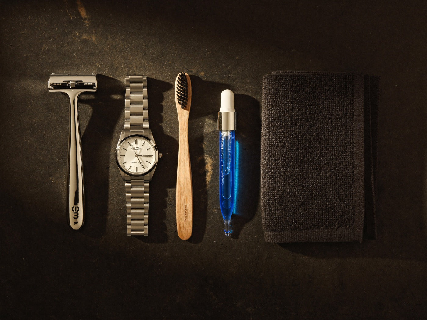 Overhead flat-lay of a razor, toothbrush, watch, towel, and glass dropper of blue serum on dark stone