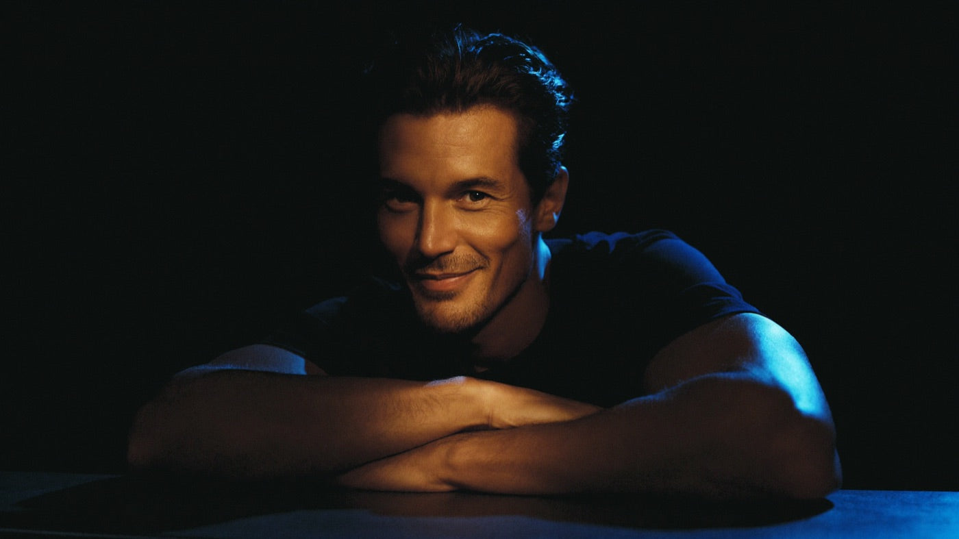 Confident man with a slight smile leaning on a dark counter in warm key light with a cobalt-blue rim accent