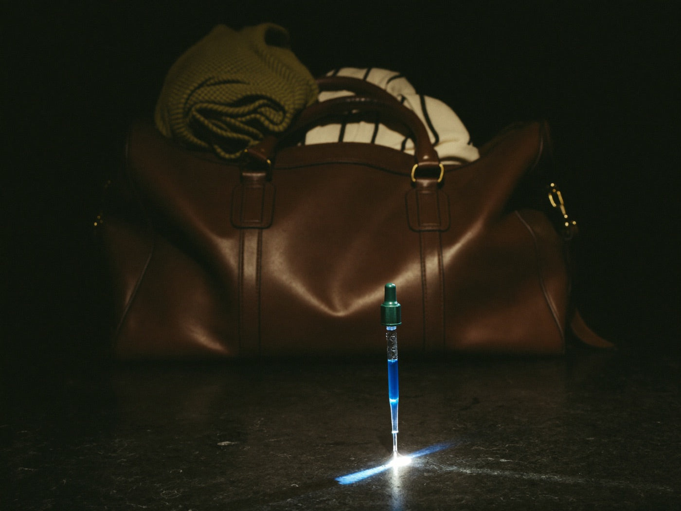 Packed leather travel bag in deep shadow behind a small glass dropper of blue serum standing in a beam of light