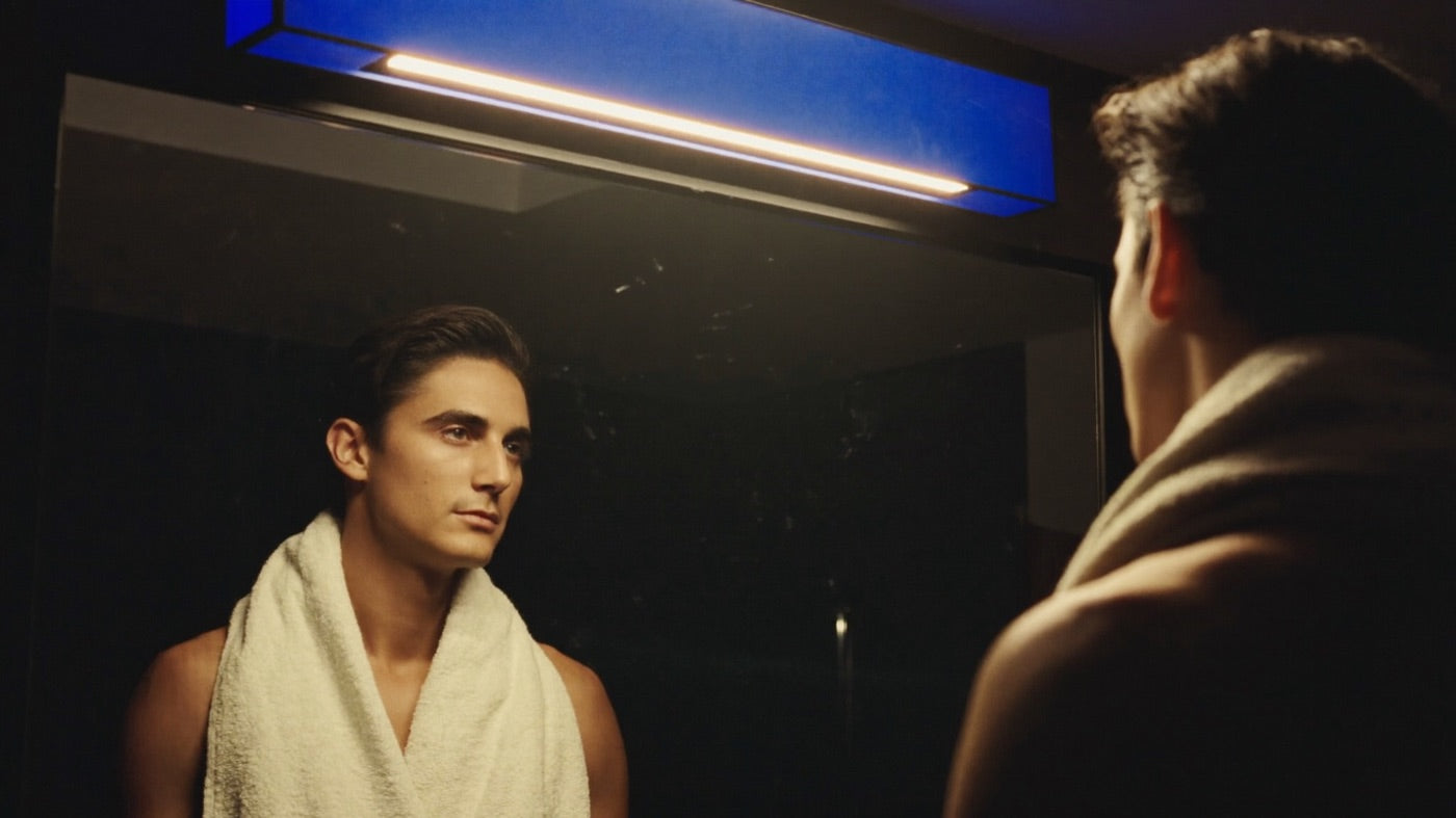 Man with a towel around his neck looking at himself in the bathroom mirror under moody light