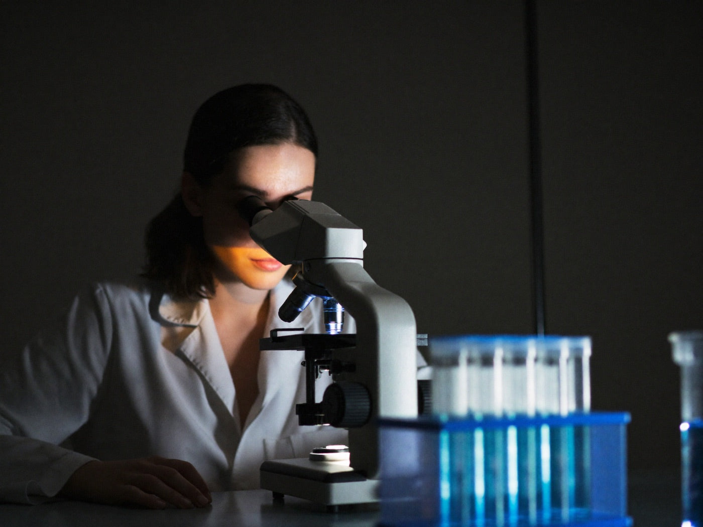 Researcher in a lab coat at a microscope in a dim laboratory, vials of blue liquid in the foreground