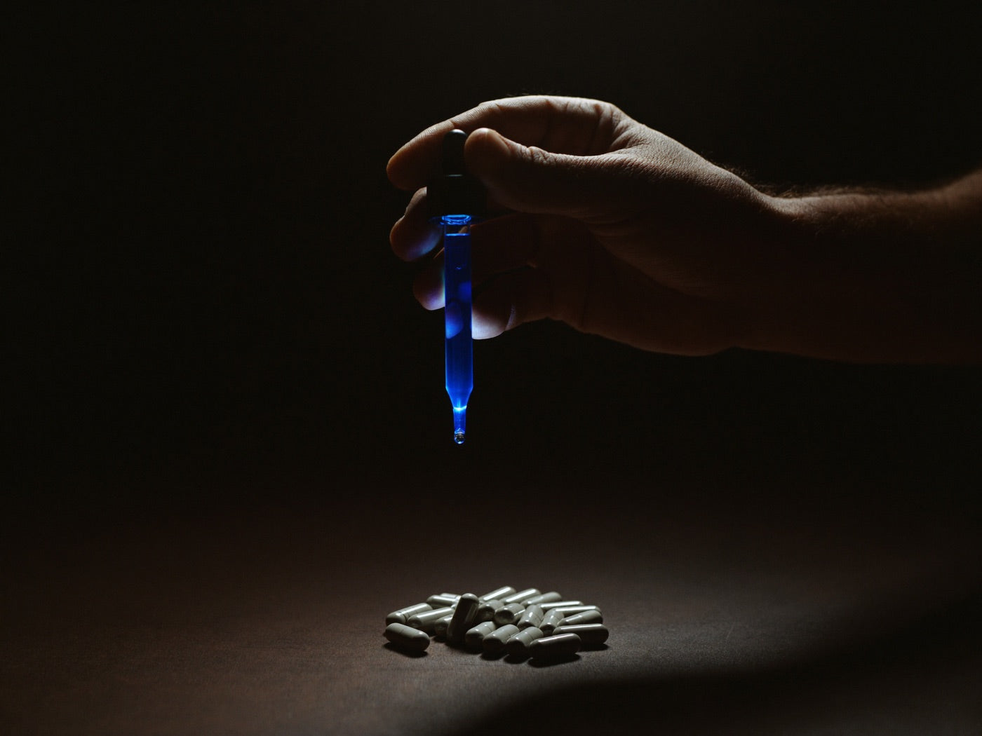 Hand holding a backlit glass dropper of glowing blue serum above a pile of gray capsules left in shadow