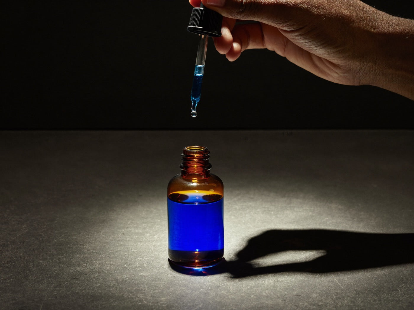 Hand holding a glass pipette above an open amber bottle of blue serum with a single falling drop