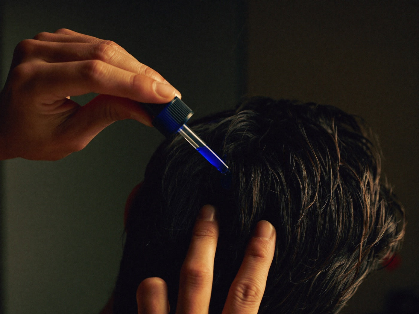 Man applying a drop of blue serum from a glass pipette dropper into his hair