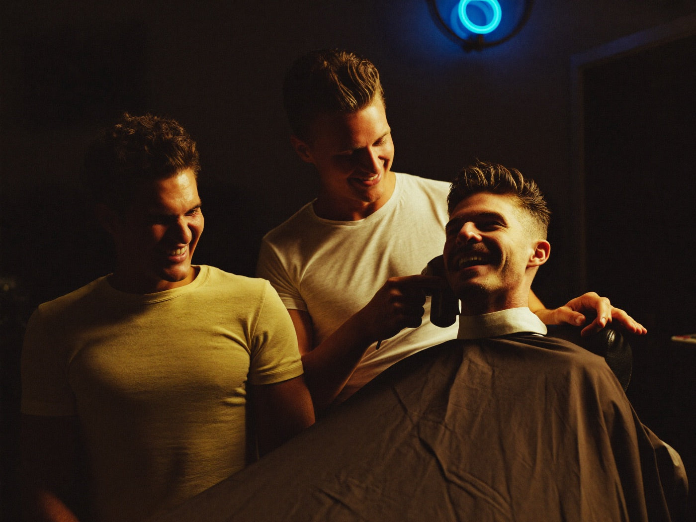 Three men laughing together in a moody barbershop, one in the chair with a fresh haircut