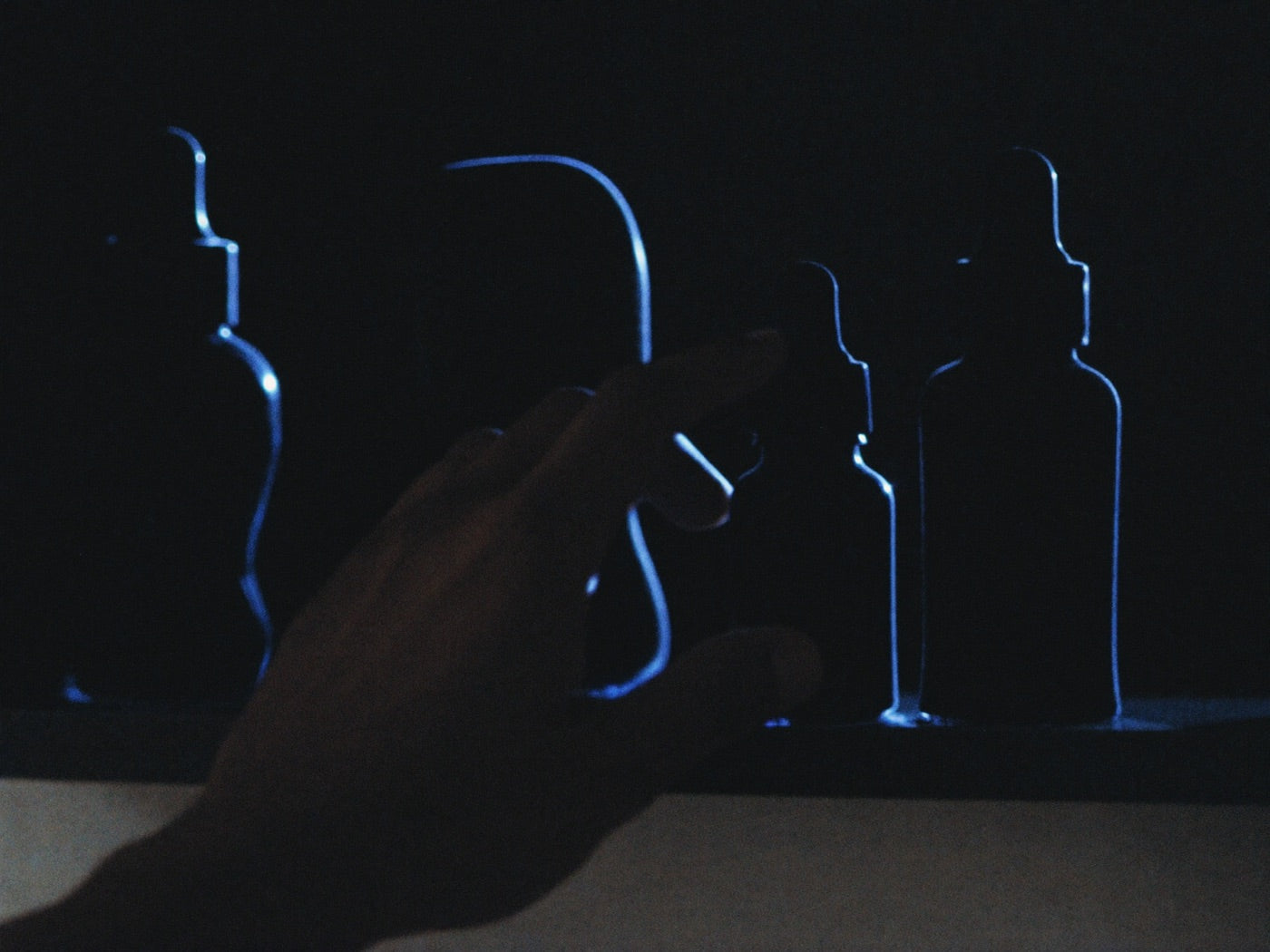 Hand reaching for a small blue glass dropper bottle glowing on a dark cabinet shelf among shadowed products