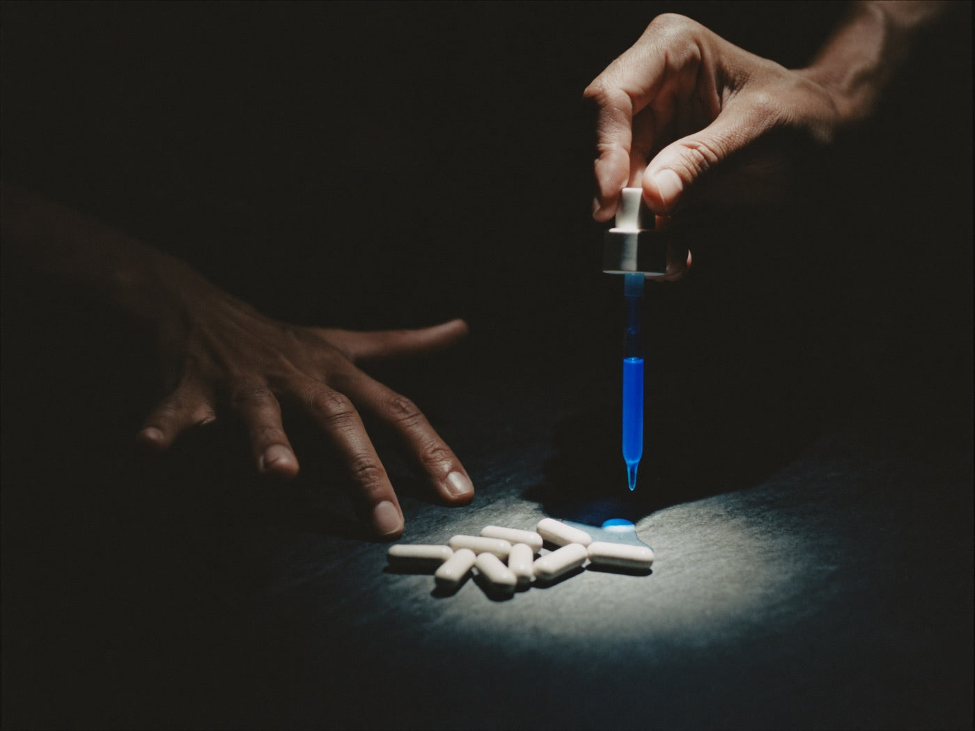 Hand sweeping gray capsules into shadow while another hand lifts a glass dropper of blue serum into the light