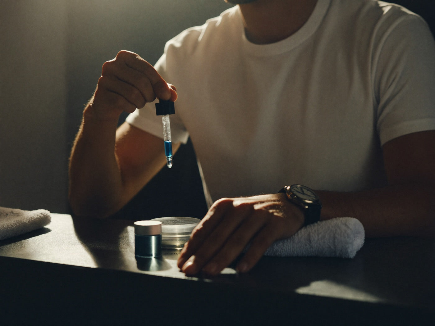 Man at his bathroom counter in morning light lifting a glass dropper of blue serum, wristwatch and towel beside him